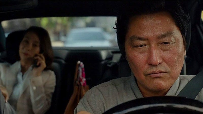 Still shot from the 2019 movie, Parasite. A man in a driver’s seat grips the steering wheel tightly, his face tense and eyes fixed ahead. In the backseat, a woman smiles brightly, looking out the window at the rain-soaked city. The contrast between their expressions is stark—his marked by quiet frustration, hers by carefree delight.