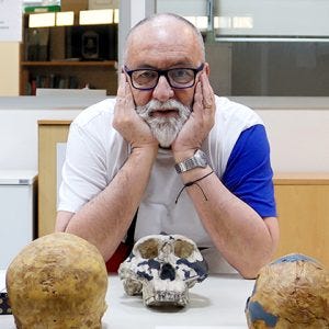 Um homem branco de óculos, parcos cabelos grisalhos nos cantos da cabeça e barba cheia branca senta-se com os ombros apoiados sobre uma mesa e as mãos apoiadas nos lados do rosto. Ele olha para a câmera. Em sua frente, um crânio apoiado sobre a mesa também olha para a câmera.