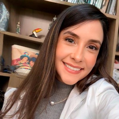Mulher sorrindo para a câmera, usando blusa de gola alta cinza e casaco branco. Ela tem cabelos longos e castanhos. Ao fundo, uma estante com livros e objetos decorativos Mulher sorrindo para a câmera, usando blusa de gola alta cinza e casaco branco. Ela tem cabelos longos e castanhos. Ao fundo, uma estante com livros e objetos decorativos
