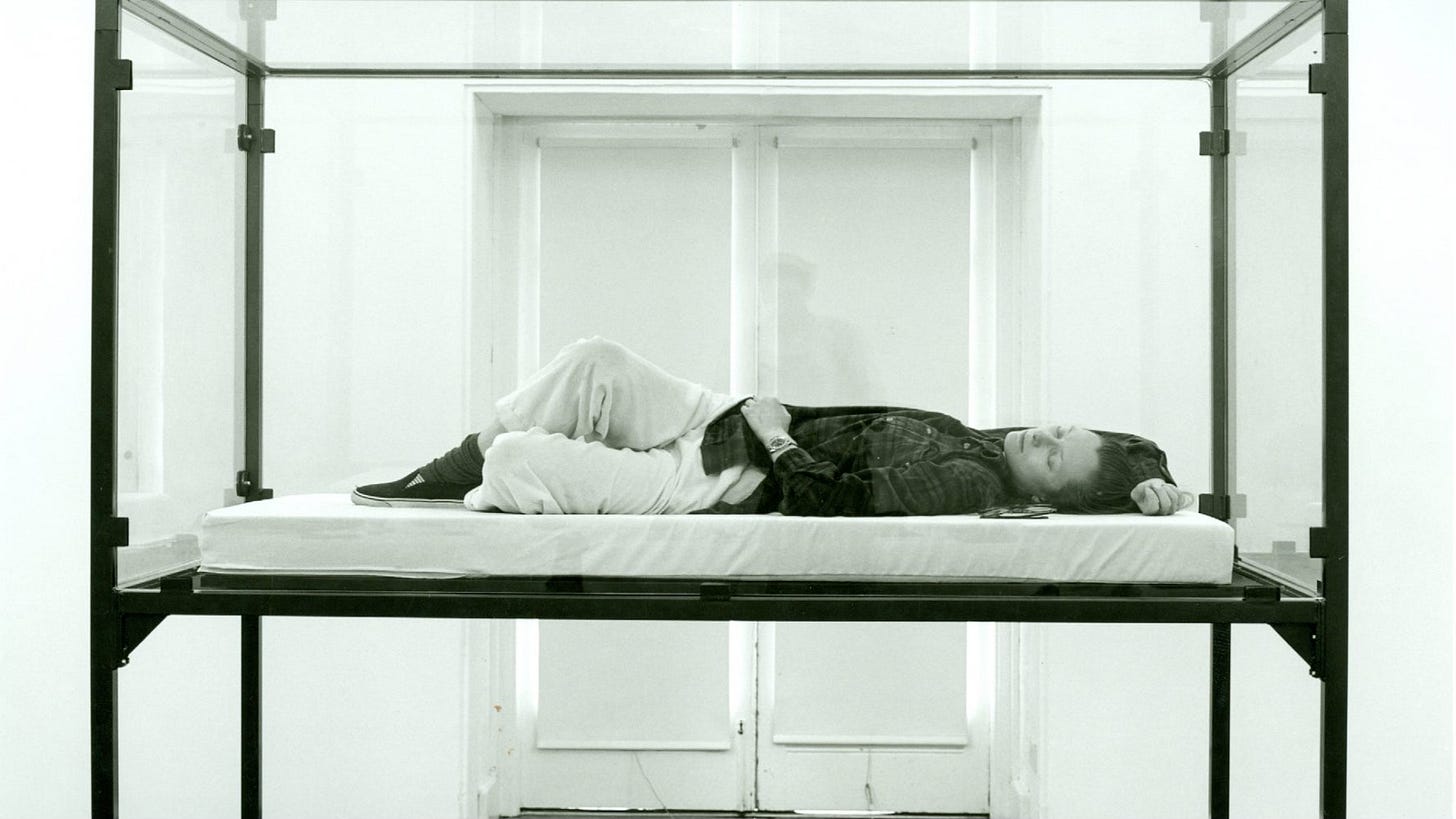 Black and white photograph from 1995 of a young Tilda Swinton, lying down on a mattress enclosed in a glass box elevated on metal legs. She wears a plaid shirt, light jeans, bunched-up socks and slip-on sneakers, and is in a state of repose on her back with her legs folded and one hand beneath her head