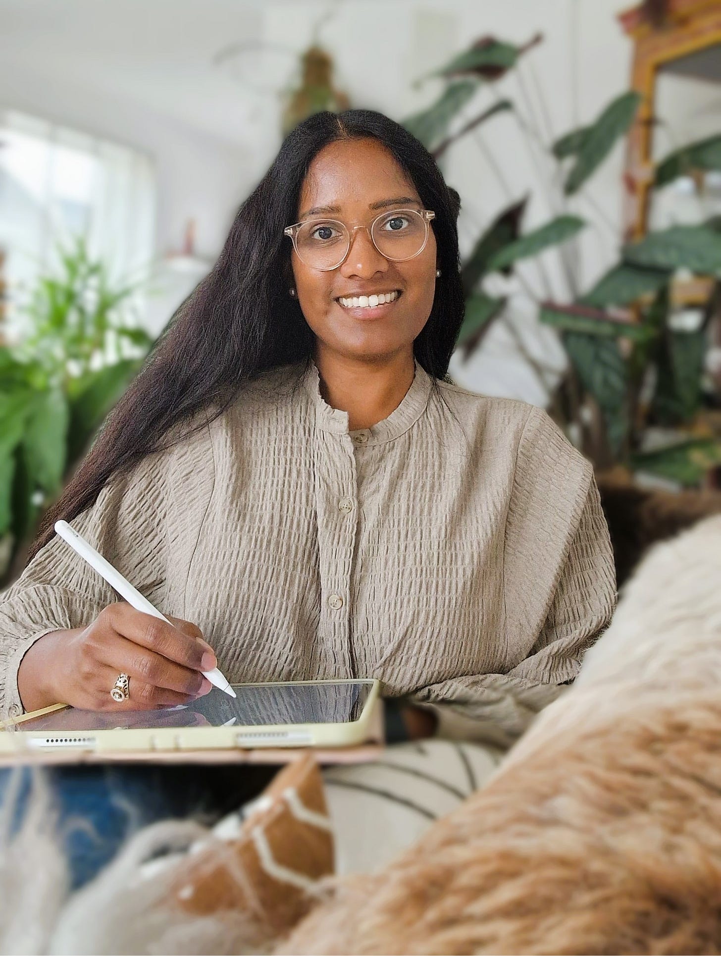 A woman with dark wavy hair and glasses smiling whilst holding an apple pencil and ipad.
