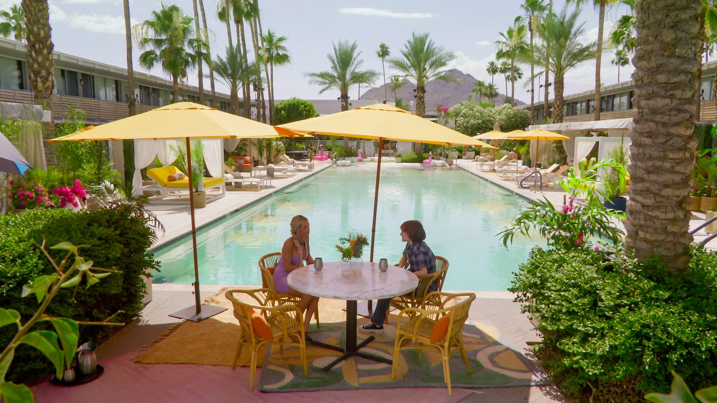 Two people sit at an outdoor table in front of the pool at a motel, with multiple palm trees and a desert mountain in the distance, in &#039;Sneaky Links.&#039;
