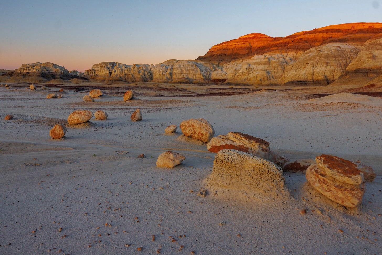 Evening light on round, eroded boulders scattered across a pale clay flat near striped badlands cliffs in the Bisti/De-Na-Zin Wilderness.