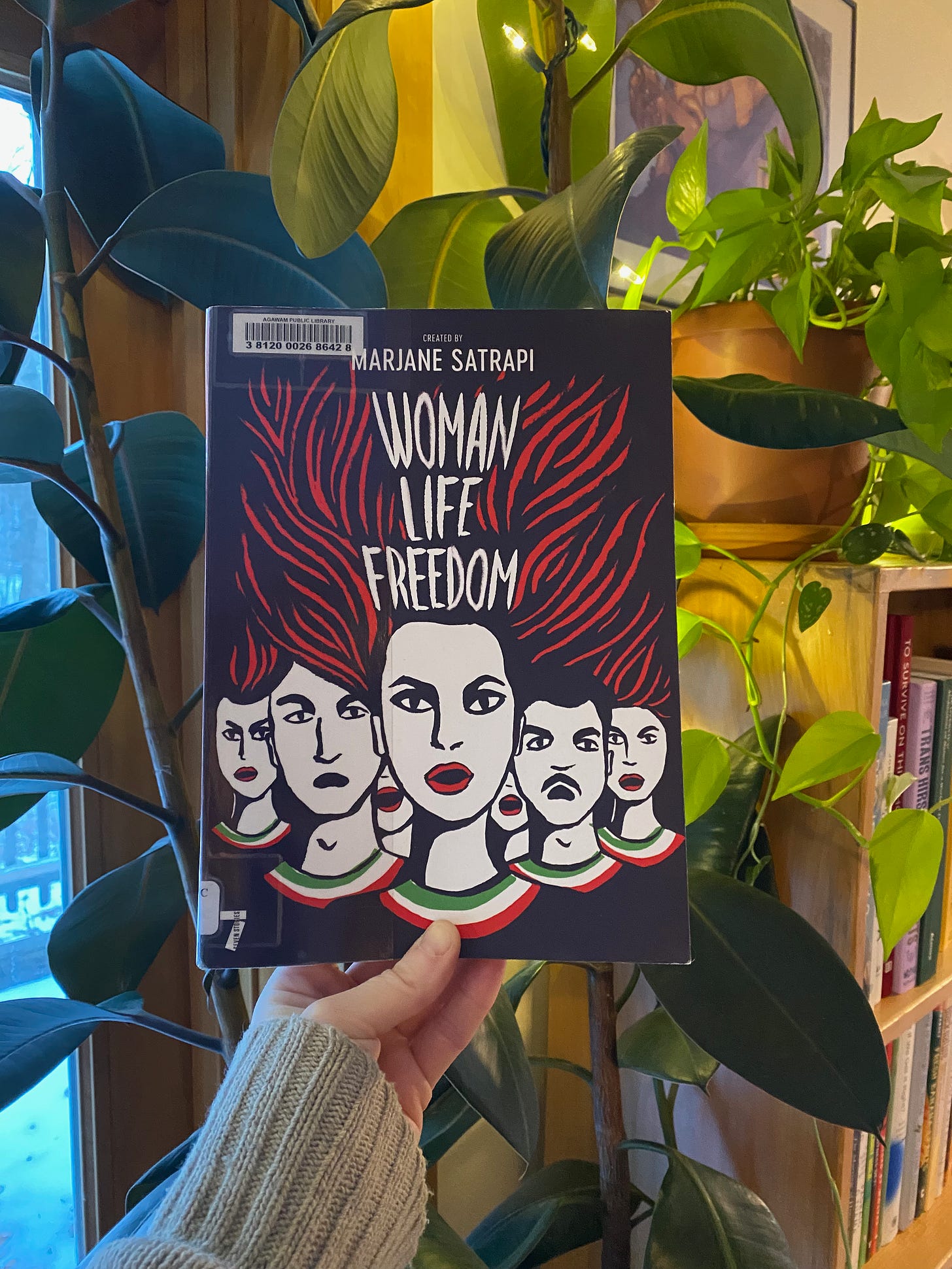 I’m holding up Woman, Life, Freedom in front of some plants next to a bookshelf.