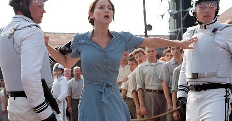 Print colorido da cena em que Katniss se voluntaria como tributo. Ela está com um vestido azul claro, cabelo preso em um coque, se enfiando entre os guardas vestidos de branco e gritando que é voluntária. Atrás, varias filas de homens vestidos com roupa cor marrom claro, olhando assustados na direção dela.
