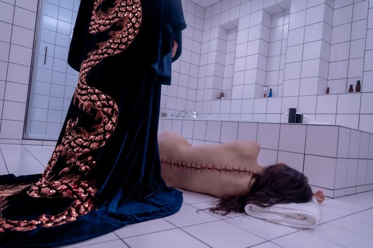 A woman in a dressing gown stands over the body of a woman with a scar down her spine.