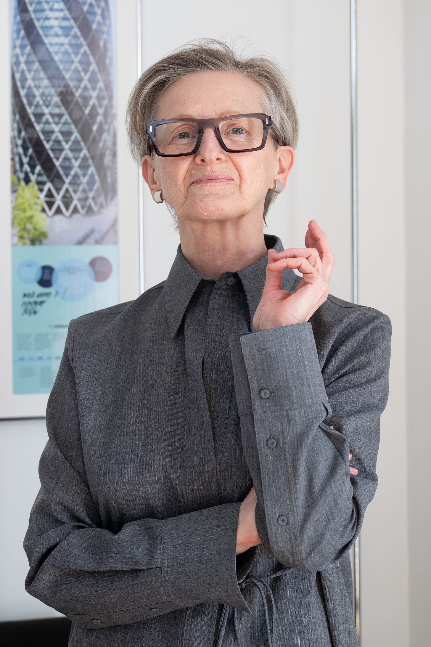 A portrait of Francesca Valli, looking thoughtfully at the camera. She is wearing dark-rimmed glasses and a grey collared shirt, standing against a neutral background with a city building visible in the distance.