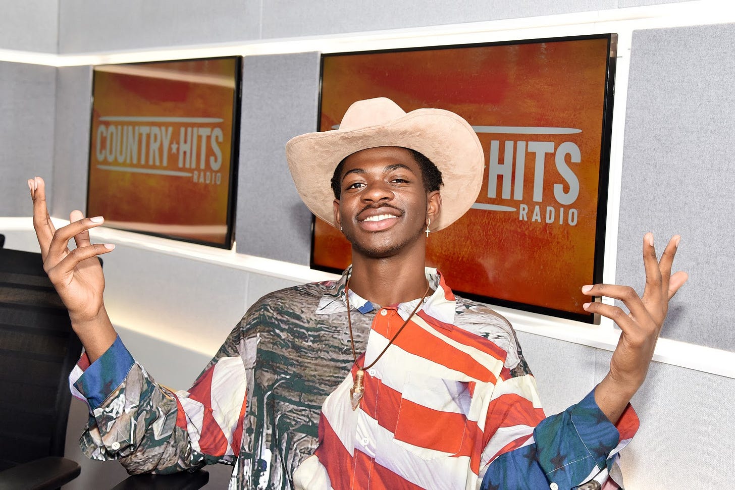 Photo of Lil Nas X wearing an American flag button down and cowboy hat, sitting in front of two televisions that display a "Country Hits Radio" emblem. Photo of Lil Nas X wearing an American flag button down and cowboy hat, sitting in front of two televisions that display a "Country Hits Radio" emblem.