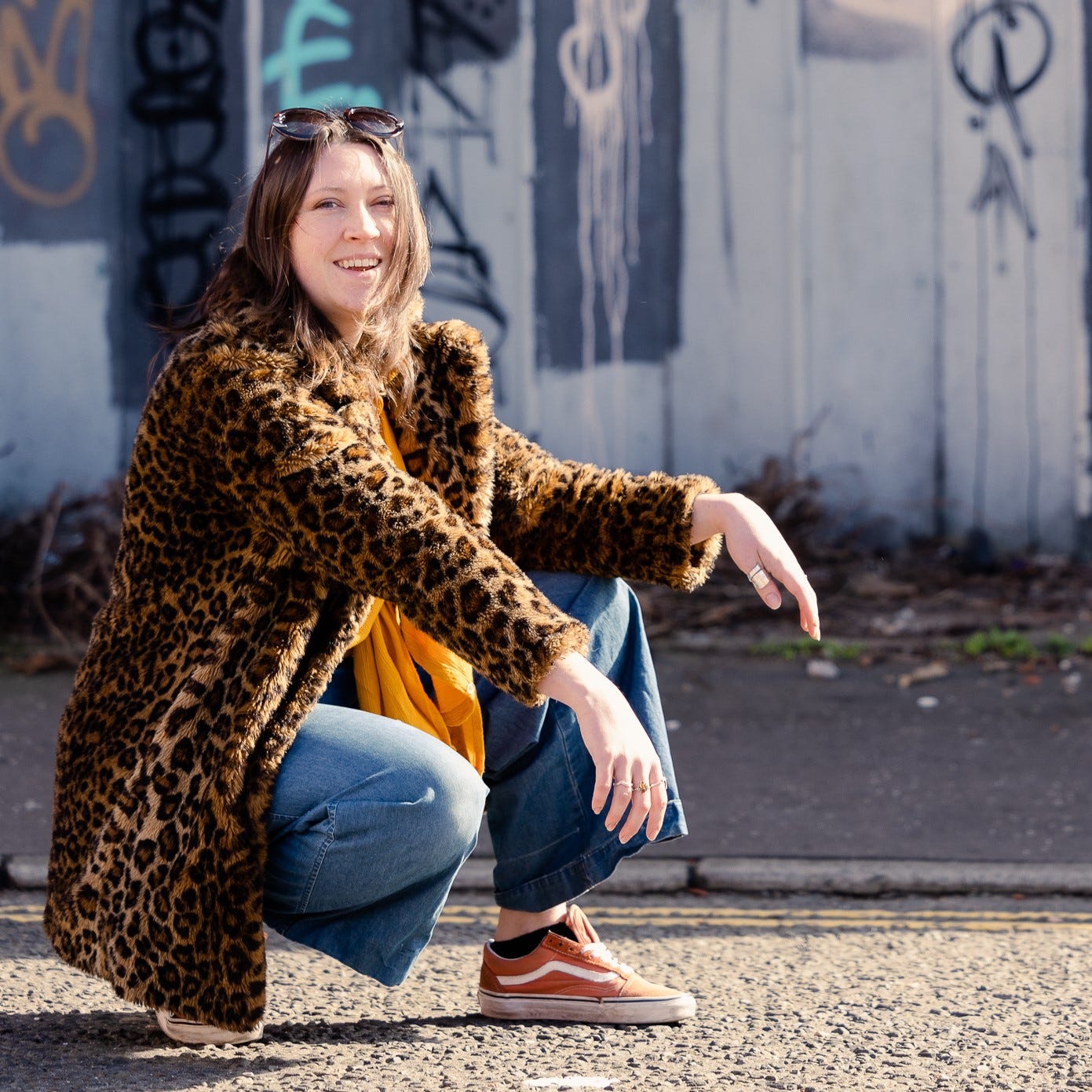 Author Natalie Jayne Clark is smiling at the camera wearing a leopard print jacket. She is crouching outside in front of graffiti on a wall.