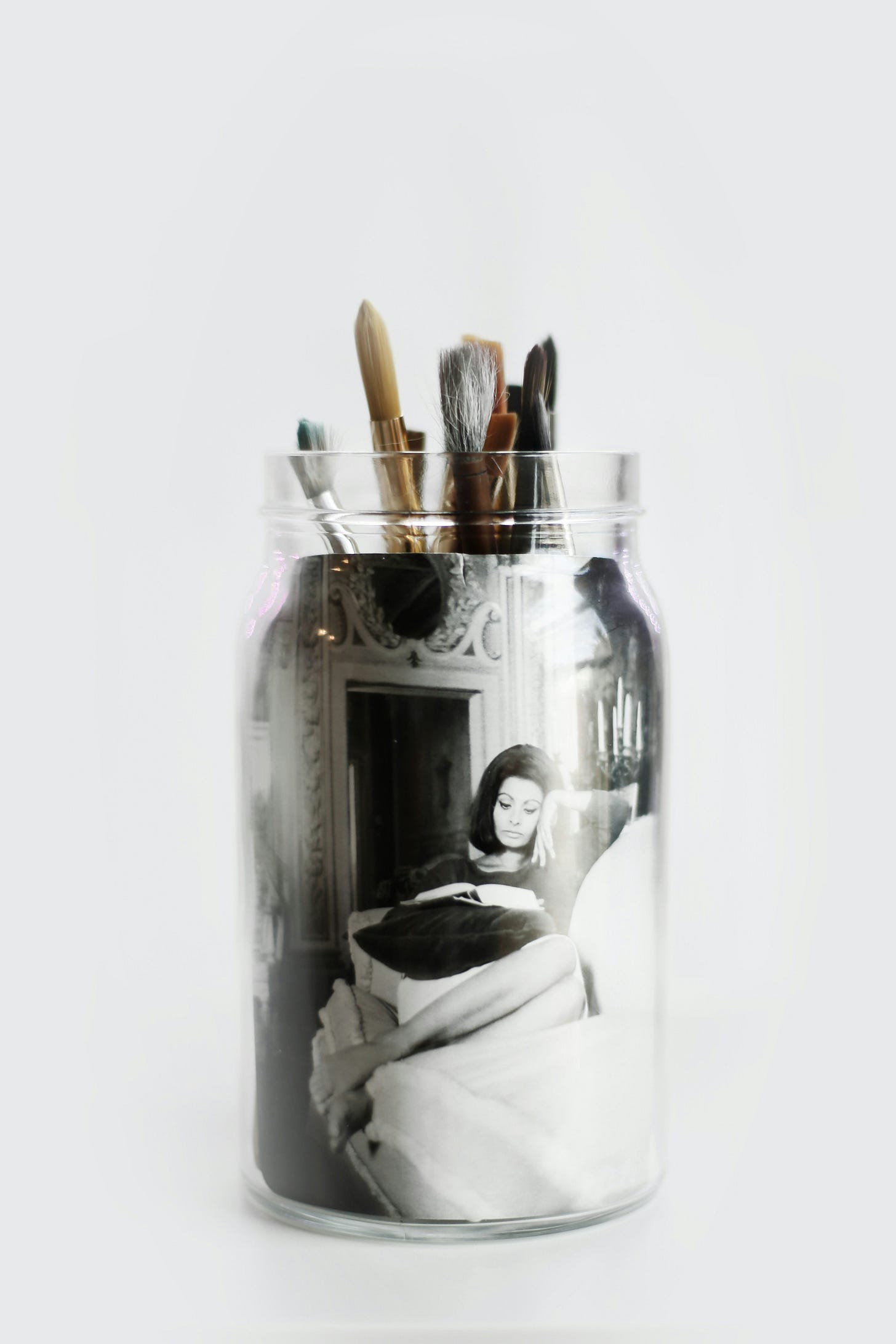 Picture of a woman is nestled inside a glass jar that has paint brushes sticking out.