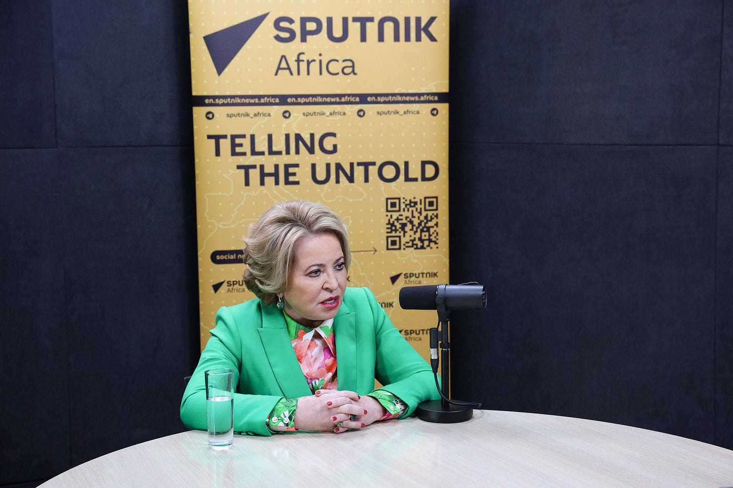 A woman in a green suit sits at a round studio desk with a microphone, in front of a backdrop reading Sputnik Africa, Telling the Untold. A woman in a green suit sits at a round studio desk with a microphone, in front of a backdrop reading Sputnik Africa, Telling the Untold.