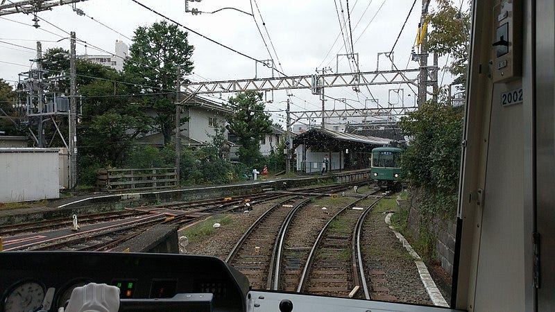 The view standing next to a train operator. Wires cross above, another train is close ahead on the tracks. The station looks small, surrounded by homes and bushy trees.