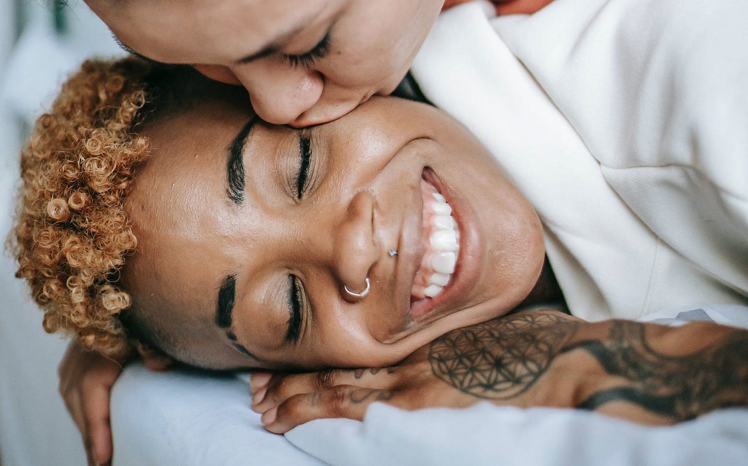A woman kisses another woman’s face while laying on a bed. The woman being kissed is smiling broadly with her eyes closed. She looks very happy.