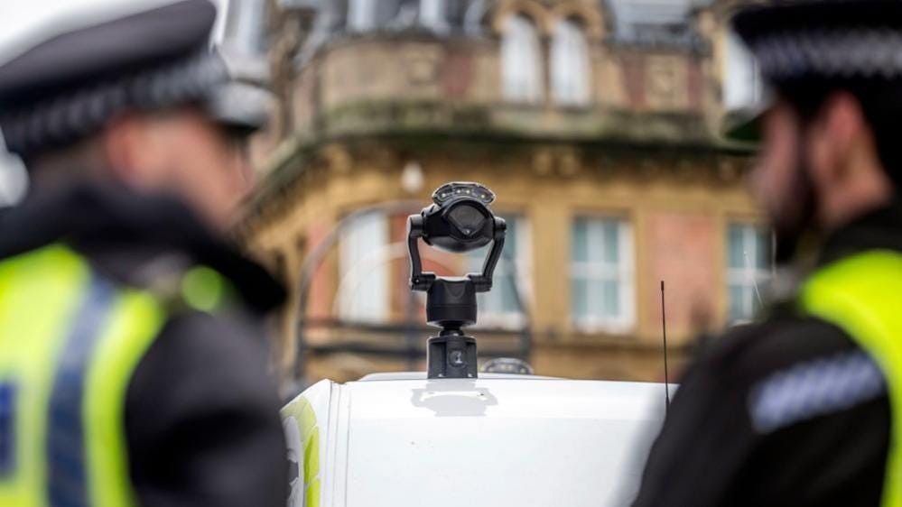 Police officers stand behind a van with a facial recognition camera on its roof