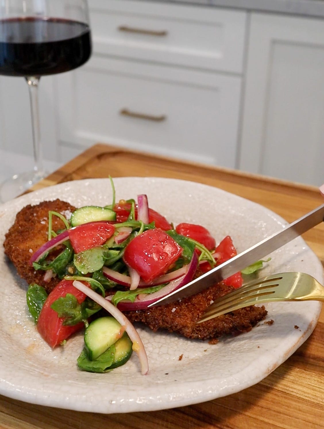 Crispy chicken Milanese topped with marinated tomato, cucumber, red onion, and arugula salad, served with a glass of red wine.