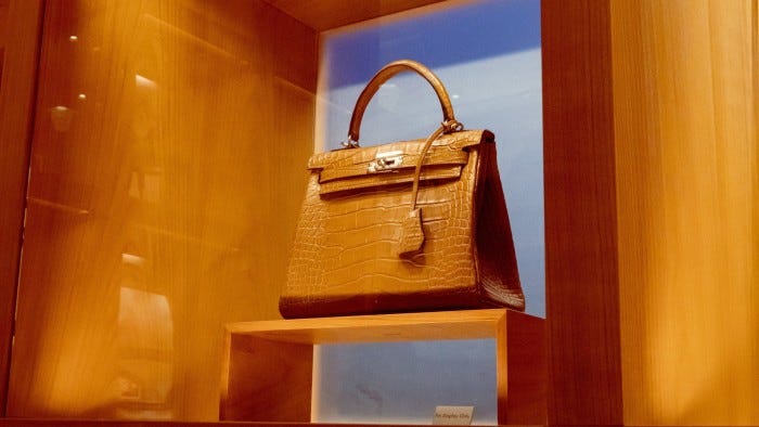 A brown Hermès Kelly bag with a crocodile pattern is displayed in a wooden case with a “For Display Only” sign.