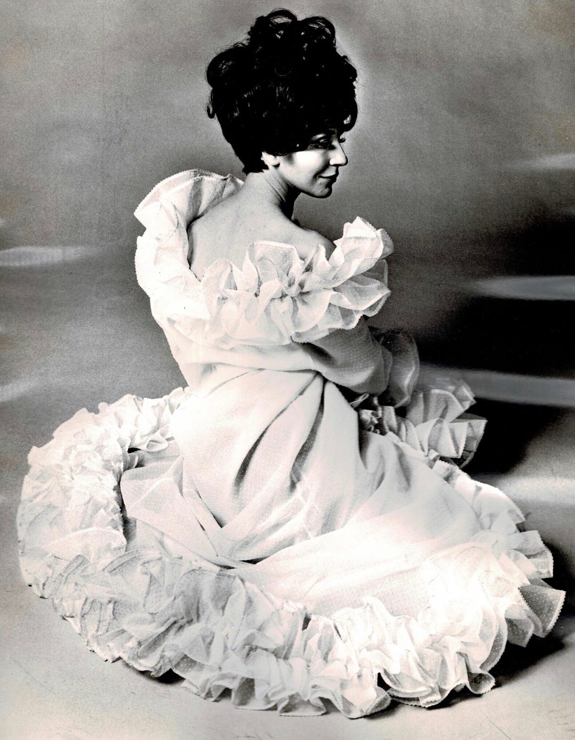 Fenella Fielding in white frilled off shoulder dress in 1966 for Harpers Bazaar Magazine wearing false eyelashes and  high hair in a bouffant style . 