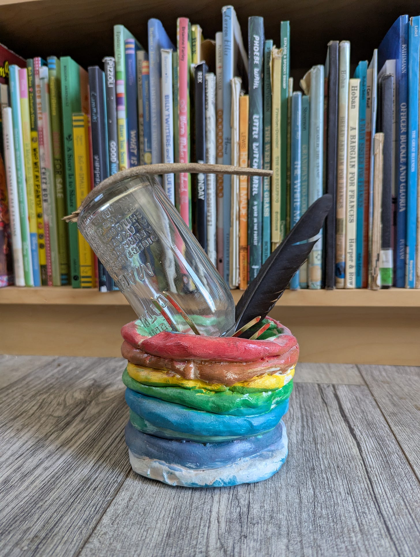 A photo of what could ostensibly be called a sculpture. It features a rainbow-colored clay coil pot (made by one of my children, yes), an antique bottle, a piece of wood, a crow feather, and some metal hair pins.