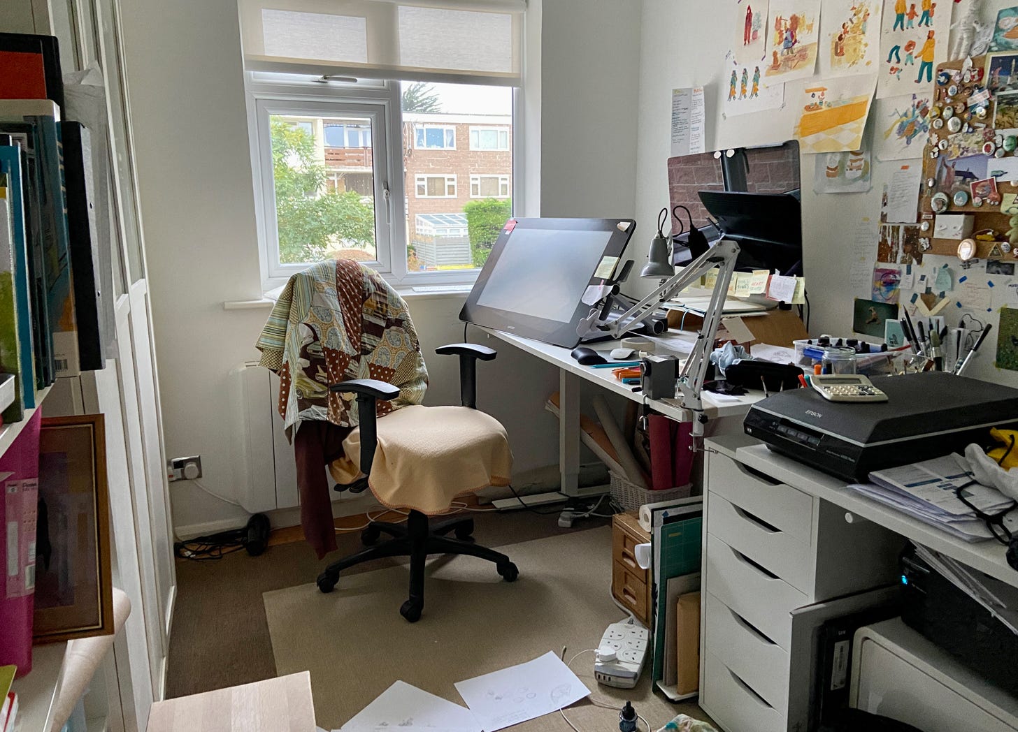 A messy cluttered desk in a small suburban room. There are artists materials and paper on the desk and floor.
