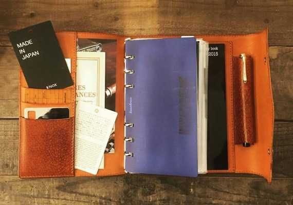 This may contain: an open leather notebook with two pens and a notepad in it on a wooden table This may contain: an open leather notebook with two pens and a notepad in it on a wooden table