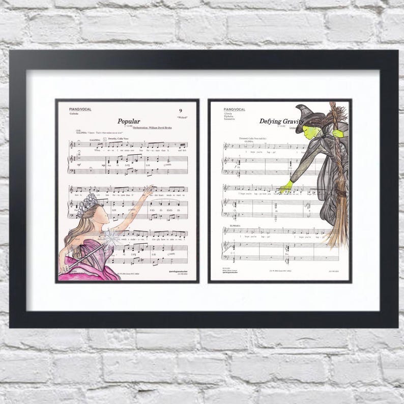 May include: A framed print of two musical scores. The left score is titled 'Popular' and features a pink and white illustration of a woman in a dress. The right score is titled 'Defying Gravity' and features a green and black illustration of a witch on a broomstick.