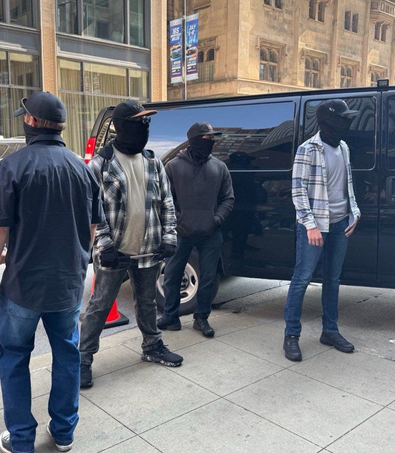 Five individuals wearing dark clothing and black masks covering their faces stand on a sidewalk. They are positioned near a black van parked on the street. The background includes a large building with windows and banners, and the ground is paved with tiles.