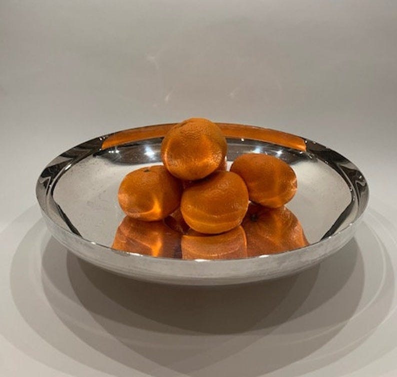 May include: A polished silver bowl holds a small pile of bright orange mandarin oranges. The round bowl is shallow and wide, reflecting the oranges and the surrounding light. The fruit is centered in the bowl, creating a visually appealing arrangement. The metallic sheen of the bowl enhances the vibrancy of the oranges.