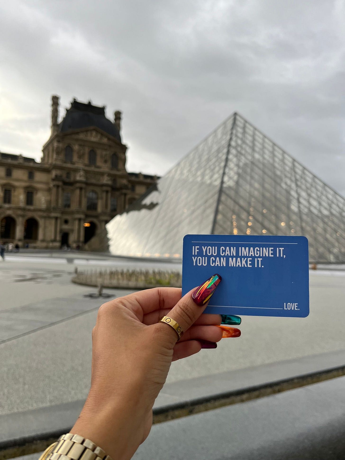 Kiana's hand holding up an affirmation card that says "If you can imagine it you can make it" in front of the Louvre pyramids