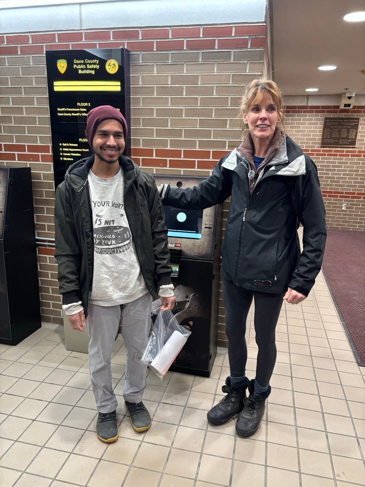 Two people smiling in the lobby of the Dane County Public Safety Building after a release from custody. One wears a maroon beanie and a t-shirt reading 'Your worth is not measured by your productivity.' A directory sign listing 'Bail and Release' and 'Initial Appearance Court' is visible behind them.