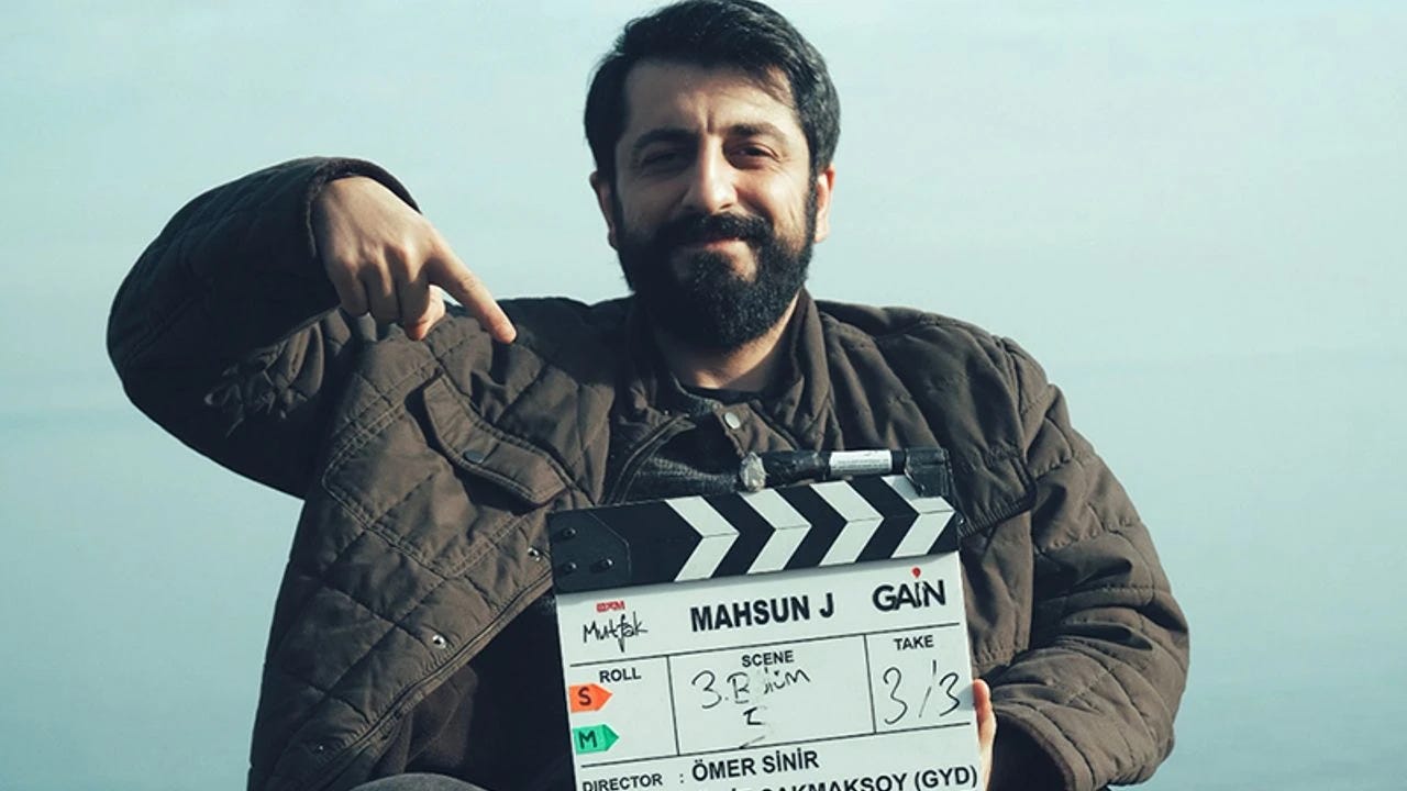 Koyu montlu bir karakter, deniz kenarında Mahsun J çekim klaketi tutuyor ve klaketi eliyle işaret ediyor; arka planda açık mavi gökyüzü var. Koyu montlu bir karakter, deniz kenarında Mahsun J çekim klaketi tutuyor ve klaketi eliyle işaret ediyor; arka planda açık mavi gökyüzü var.