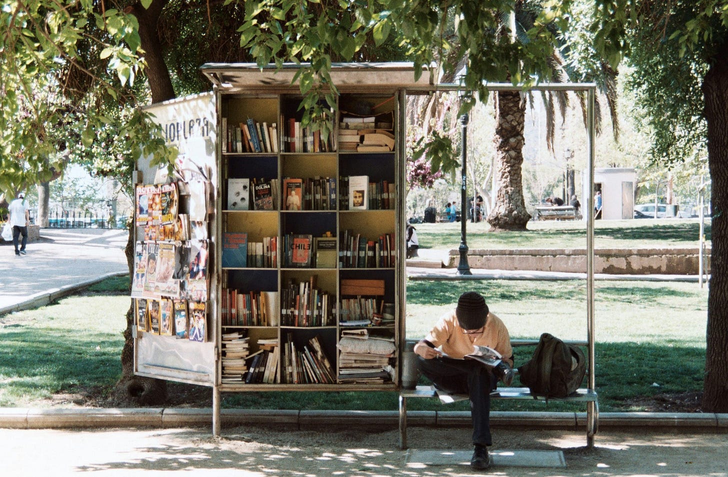 A person sits reading outdoors, on a bench in front of stocked bookshelves