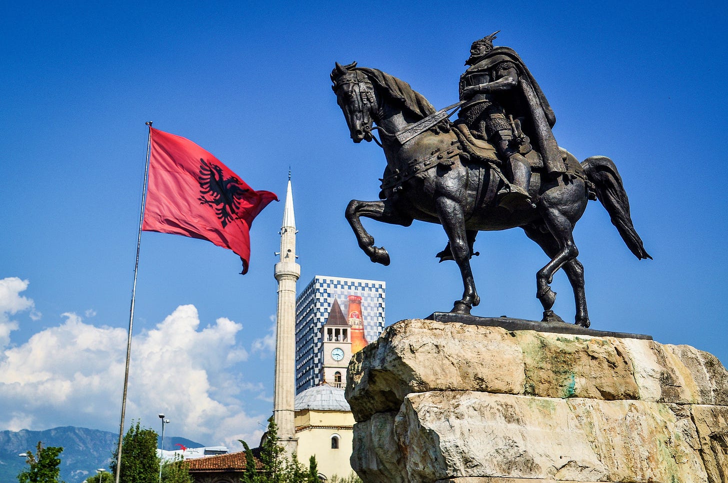 statue of man on horse overlooking albanian flag mosque behind them blue sky in background