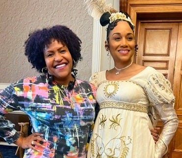 Two women offer huge happy smiles to a camera, one is author Vanessa Riley in a cream Regency gown, posing elegantly, and the other woman is producer Tia A. Smith in contemporary, colorful shirt, each have a hand on their hip Two women offer huge happy smiles to a camera, one is author Vanessa Riley in a cream Regency gown, posing elegantly, and the other woman is producer Tia A. Smith in contemporary, colorful shirt, each have a hand on their hip