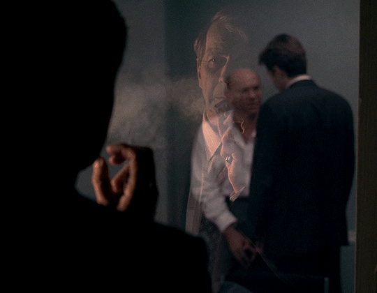 The Cigarette Smoking Man watches Mulder and Skinner talk through the window of an interview room. He is, of course, smoking a cigarette as he watches them. The Cigarette Smoking Man watches Mulder and Skinner talk through the window of an interview room. He is, of course, smoking a cigarette as he watches them.