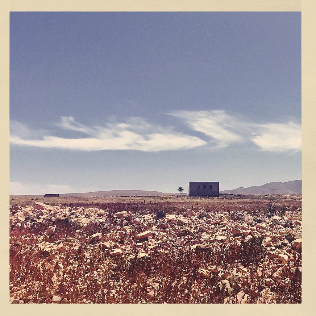 A faded Polaroid-style snapshot of a barren Fuerteventura landscape, showing rocky desert, a solitary house in the distance, and dry mountains under a clear sky