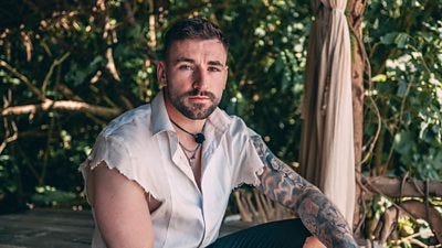 A man in a white distressed shirt with a sleeve tattoo sits in the shade on a tropical island