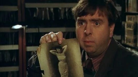 Photo of Timothy Spall in the show, holding up a black and white photo of a woman holding a provocative pose in a low-cut dress