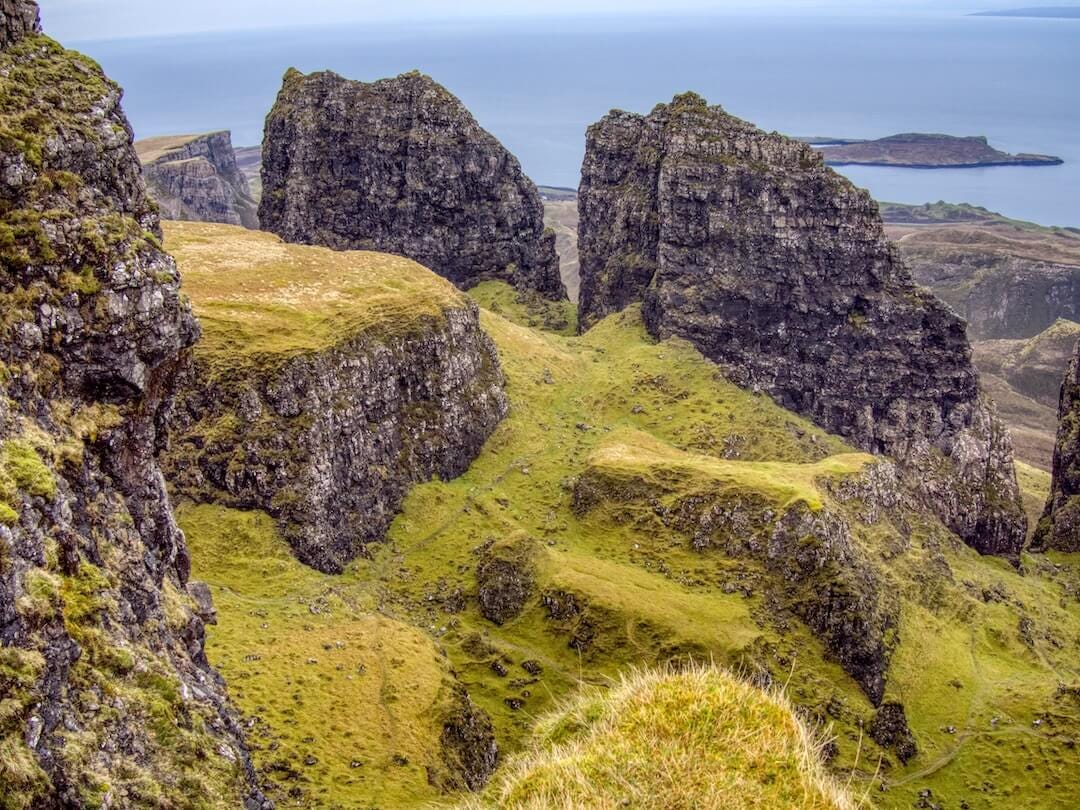 A green plateau surrounded by jagged rock formations and a blue sea in the background