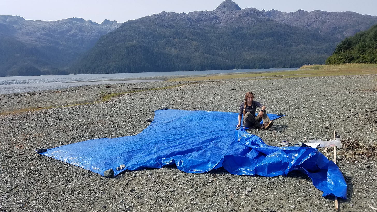 A girl with a troubled expression sits by a blue tarp on an Alaskan beach