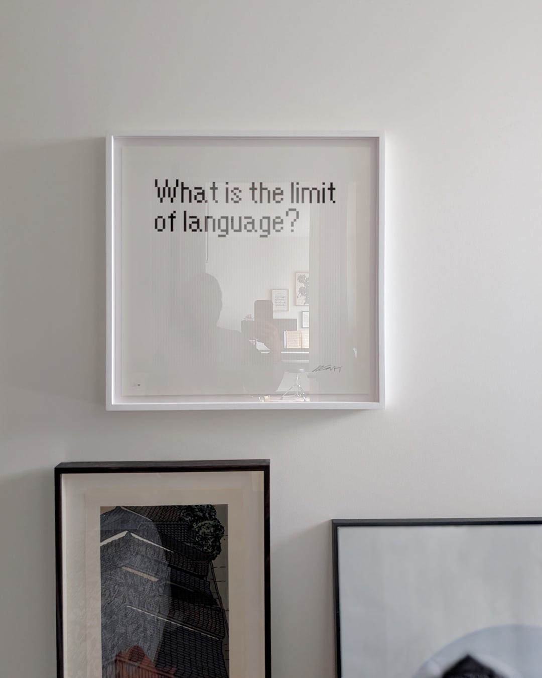 A framed print hanging on a light gray wall, showing the question "What is the limit of language?" in a pixelated, bitmap-style typeface on a white background with a subtle grid. The glazed frame reflects the silhouette of the room's interior. Below and to the sides, two additional framed works — one with a black frame, one with a dark wood frame — are partially visible leaning against the wall.