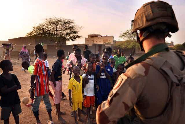 Children play soccer and stand looking at a man in military uniform