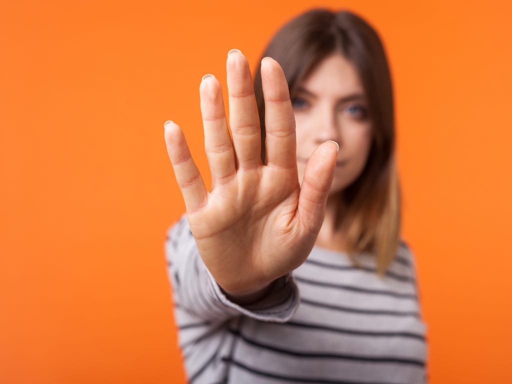 Vrouw steekt haar hand naar voren in een stopgebaar tegen een effen oranje achtergrond.