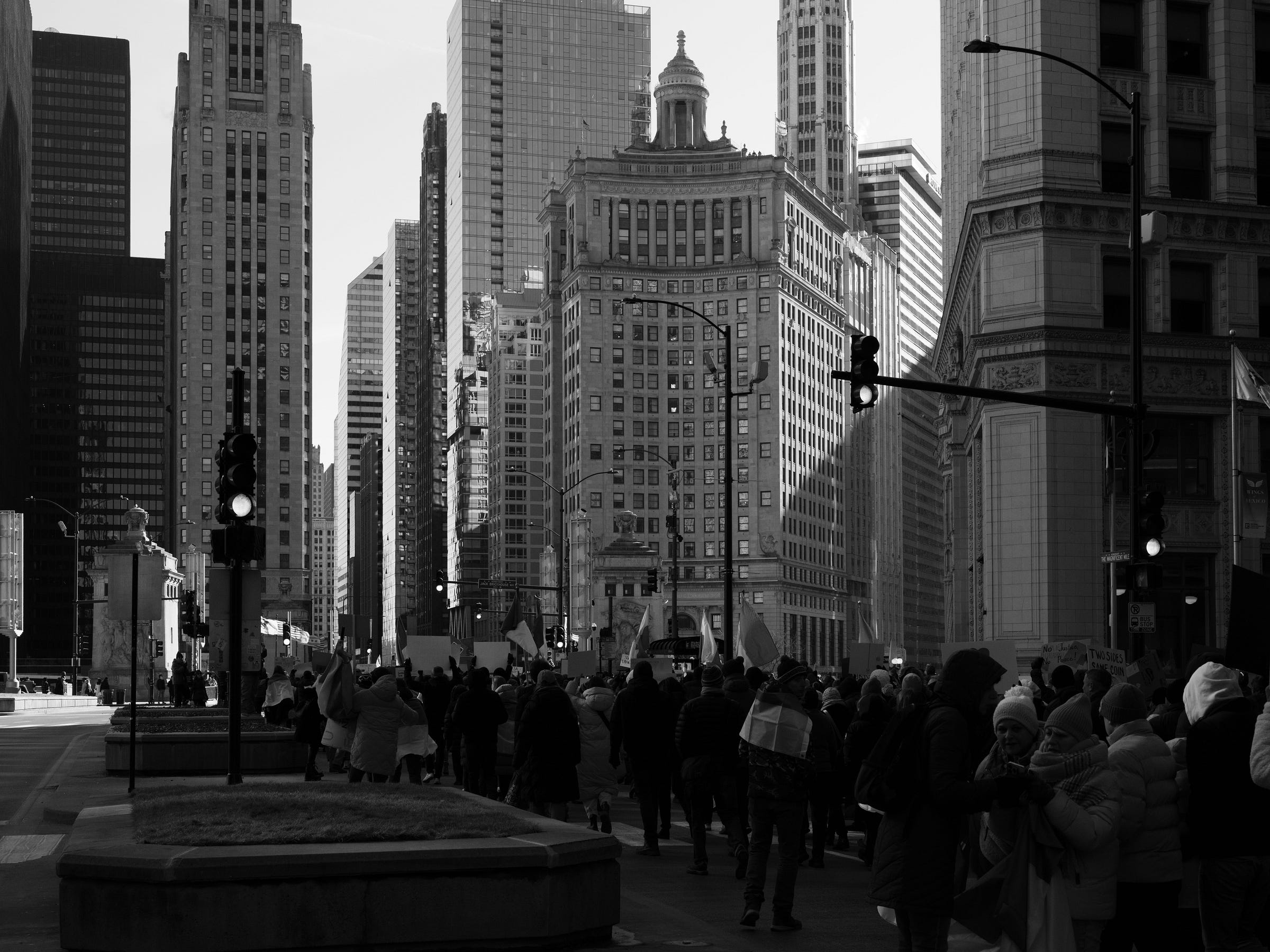 A black and white photograph captures a protest march moving through downtown Chicago, with towering skyscrapers in the background. The demonstrators, bundled in winter clothing, carry flags and signs as they walk past traffic lights and historic buildings. The contrast between the crowd and the cityscape emphasizes the scale of the movement against the urban backdrop. A black and white photograph captures a protest march moving through downtown Chicago, with towering skyscrapers in the background. The demonstrators, bundled in winter clothing, carry flags and signs as they walk past traffic lights and historic buildings. The contrast between the crowd and the cityscape emphasizes the scale of the movement against the urban backdrop.