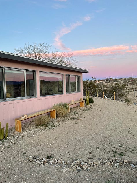 Art supplies on table with view of the desert, pinks and purples in the sky for sunset, a joshua tree at sunset, a pink house in the desert that matches the pink colors in the sky