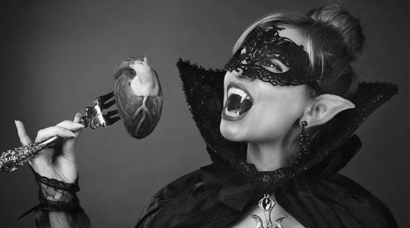 A woman in a black masquerade mask and vampire fangs prepares to eat a realistic human heart held on a silver fork.