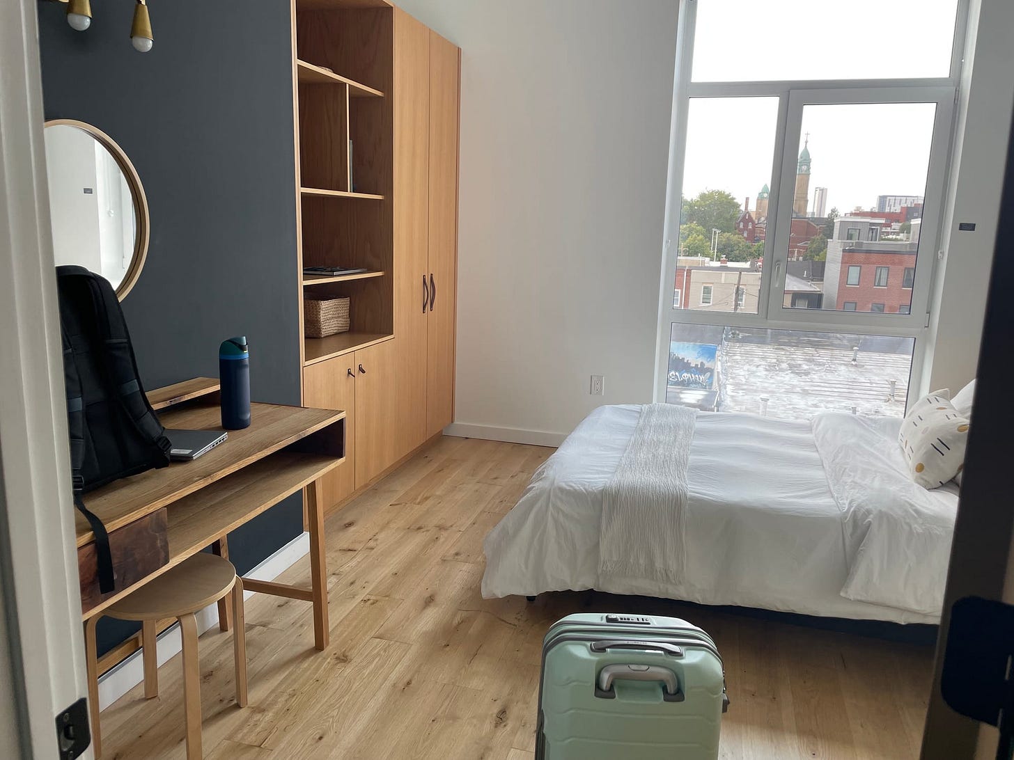 A room in a rental apartment with wood flooring and furniture, a bed with white linens, and a window looking out to a city street