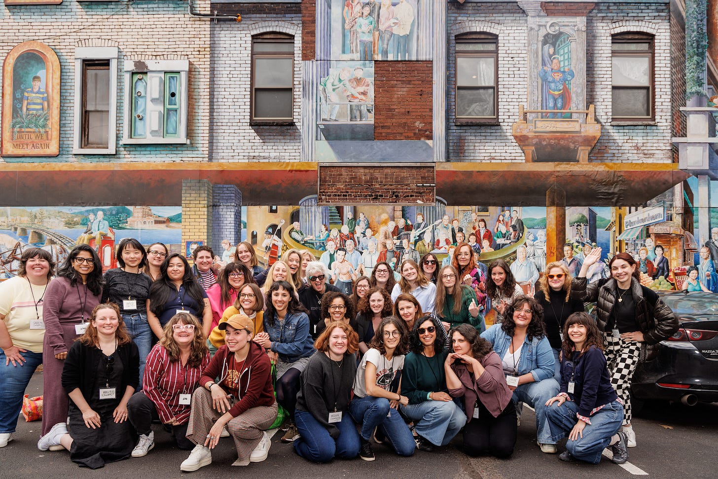 A large group of women and enby folks stand in front of a painted brick mural featuring windows and many characters A large group of women and enby folks stand in front of a painted brick mural featuring windows and many characters