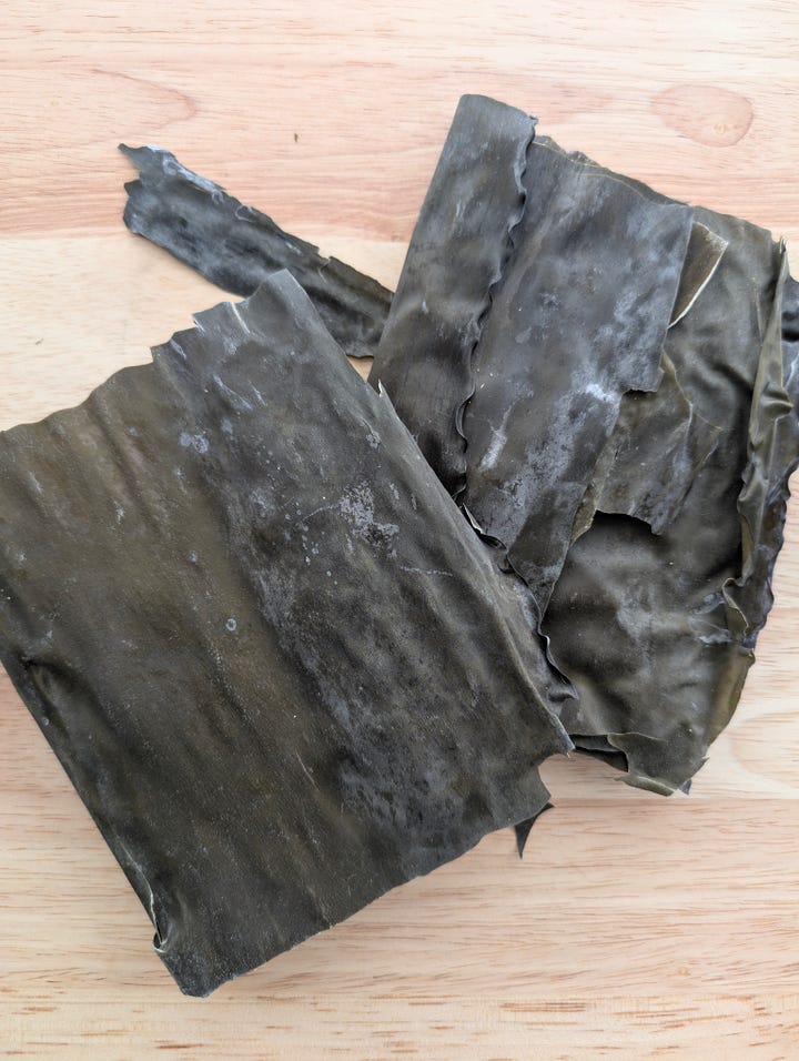 Sheets of kombu seaweed on a wooden cutting board, showing its dark, textured surface, alongside a dish of bright green pickled snap peas with kombu, served in a decorative bowl.