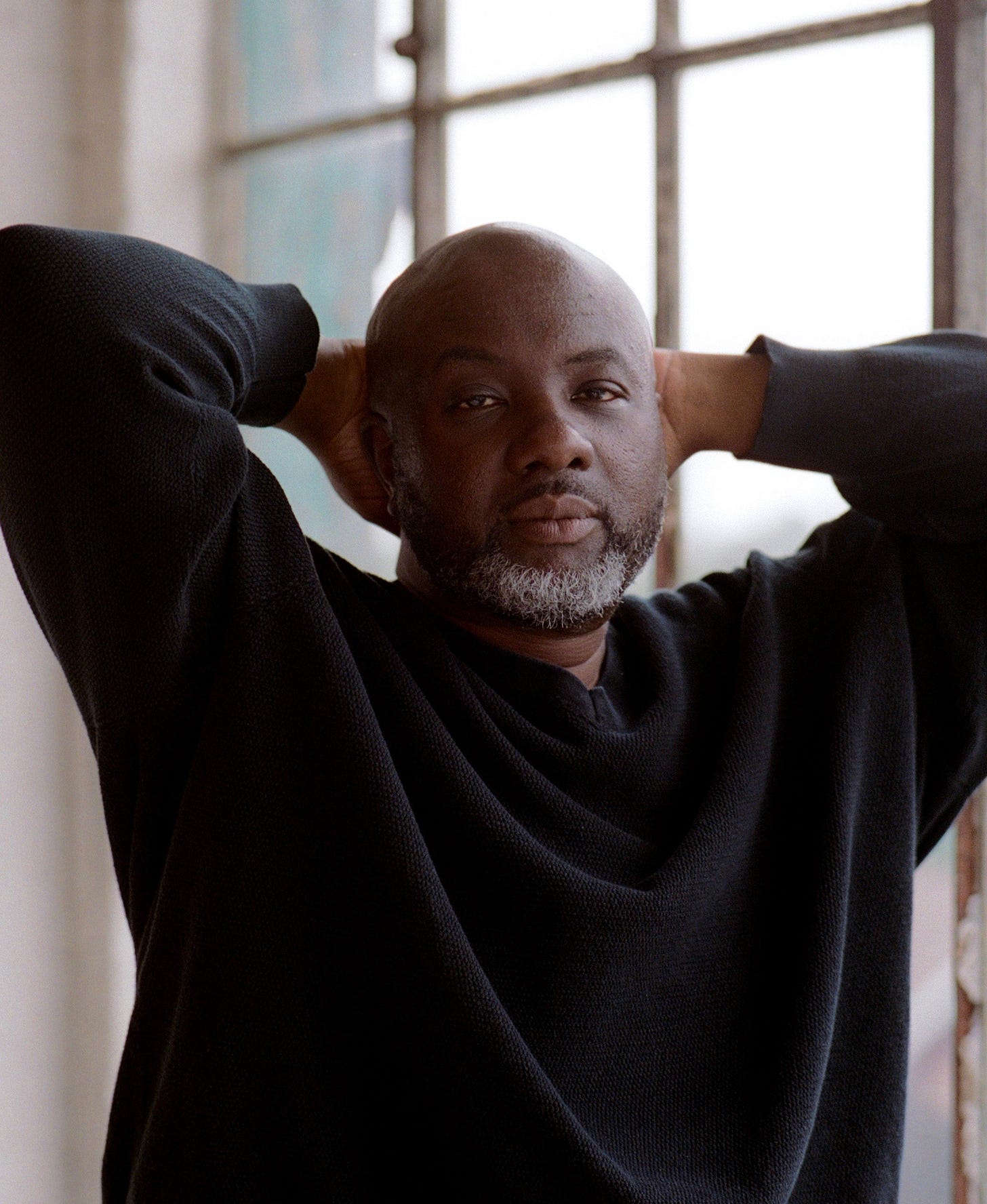 Portrait of a contemplative man with a bald head and gray beard wearing a black sweater. He's photographed with his hands behind his head against a bright window background, creating a thoughtful, relaxed pose. The natural lighting creates a warm, introspective mood.