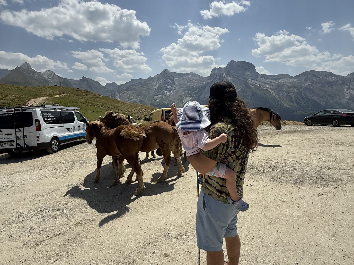 anthony holding his baby on the summit of the mountain with insane views around him and wild horses in the parking lot right next to them